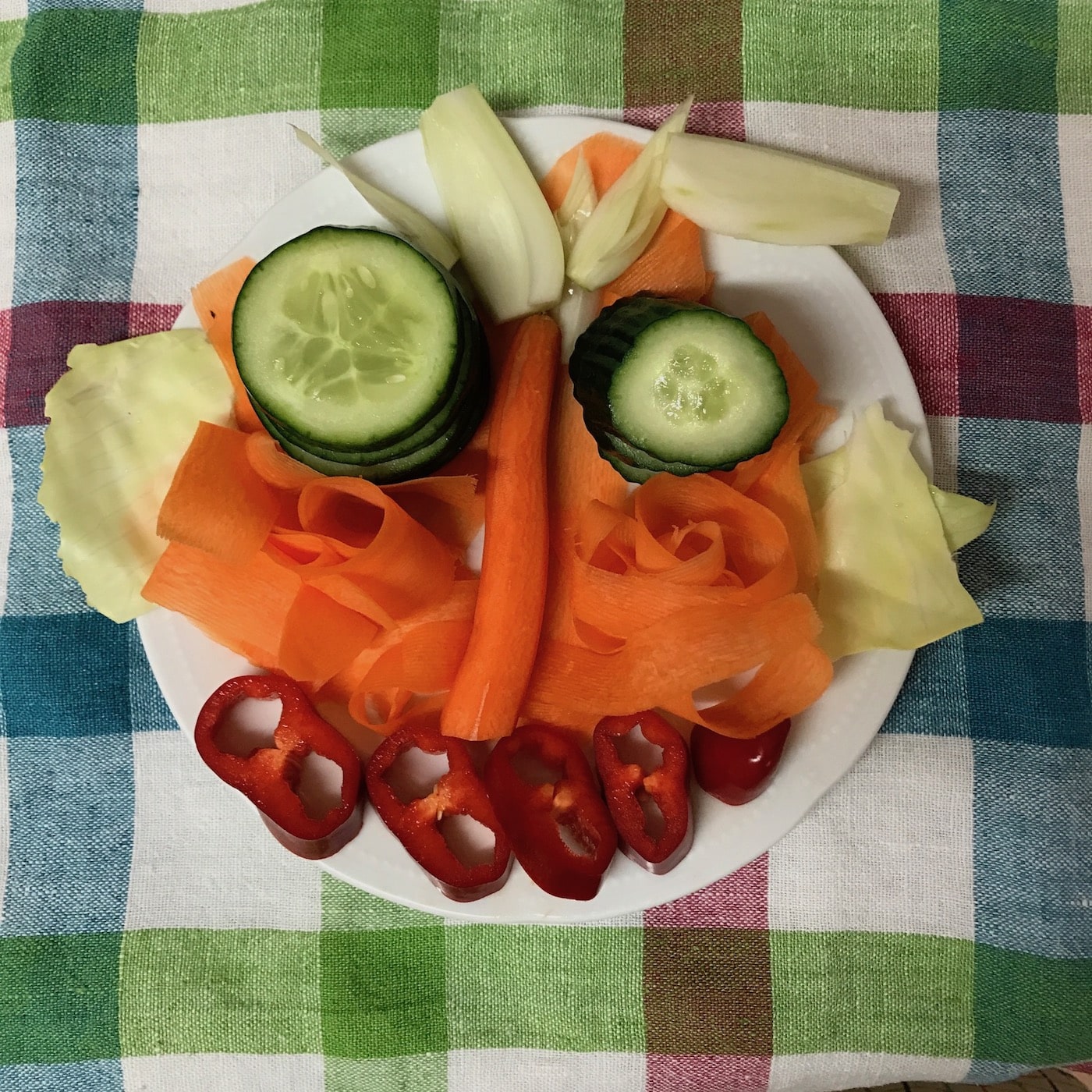 Gesicht aus Gemüse wie Gurken, Möhren, Paprika auf einem Teller angerichtet.